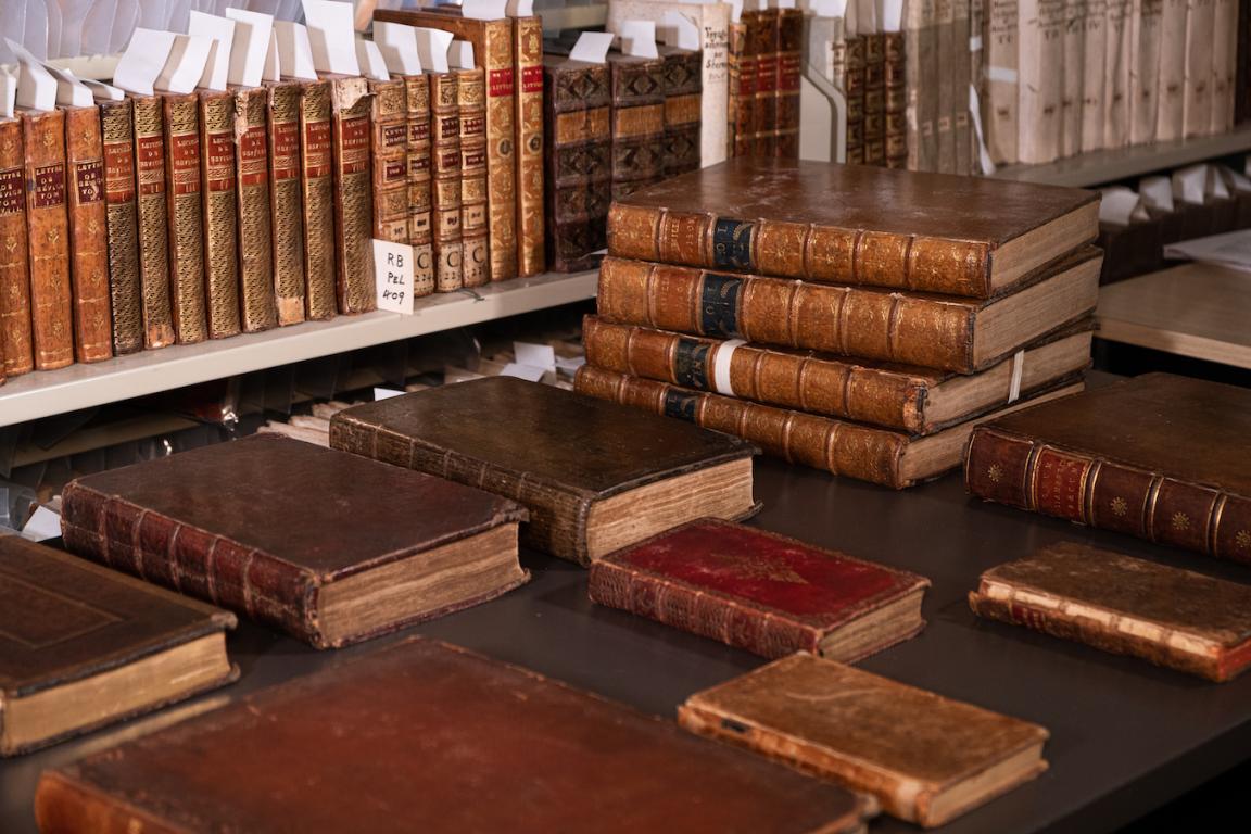 Shelf and table with dozens of old, leather-bound books, some with gold designs or writing on the spines