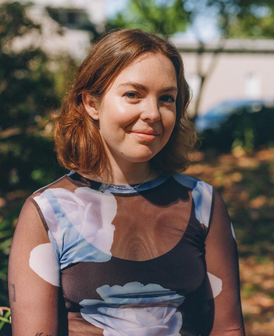 Tess has short, light brown hair and is standing outdoors wearing a dark, sheer top with large, light-coloured floral patterns and a thin necklace. The background includes greenery and a slightly blurred building and car.