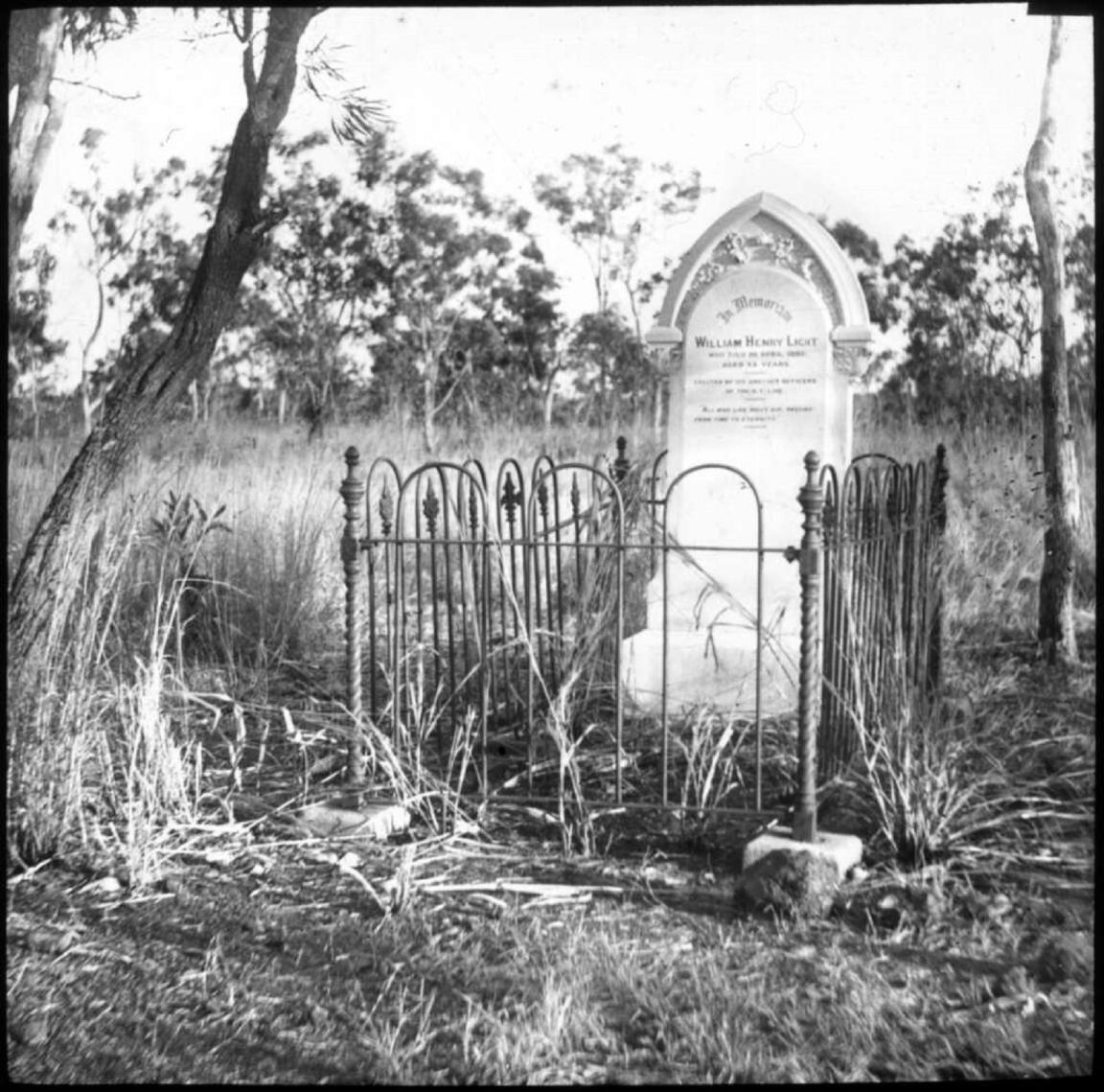 Black and white photograph of a gravestone surrounded by a wrought iron fence. The grave is situated in a grassy, rural setting with a few trees scattered around. The gravestone has an arched top and an inscription in memory of William Henry Light.