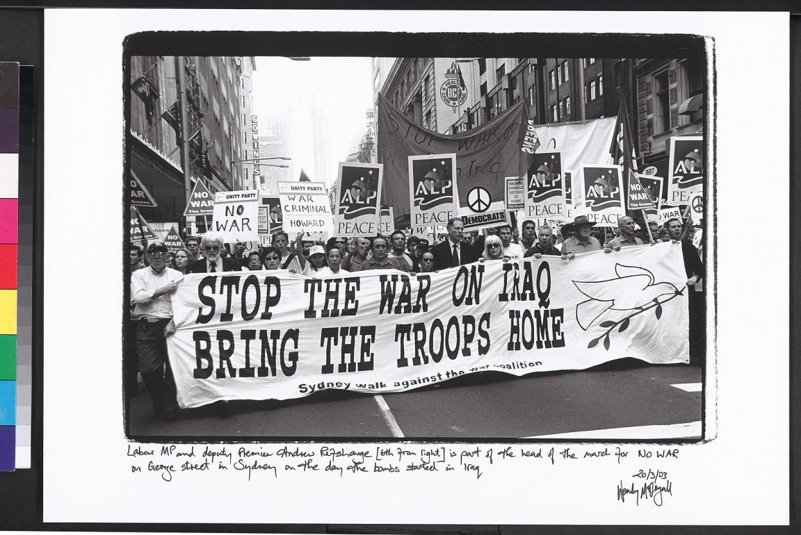A black-and-white photograph of a large protest march on George Street in Sydney, taken on the day the bombs began in Iraq. The crowd holds a large banner that reads, "STOP THE WAR ON IRAQ BRING THE TROOPS HOME," with a white dove and olive branch illustration on the right. Numerous signs are visible behind the banner, including those from the Australian Labor Party (ALP) and various anti-war groups. Signs include messages such as "NO WAR," "WAR CRIMINAL HOWARD," and "PEACE."
