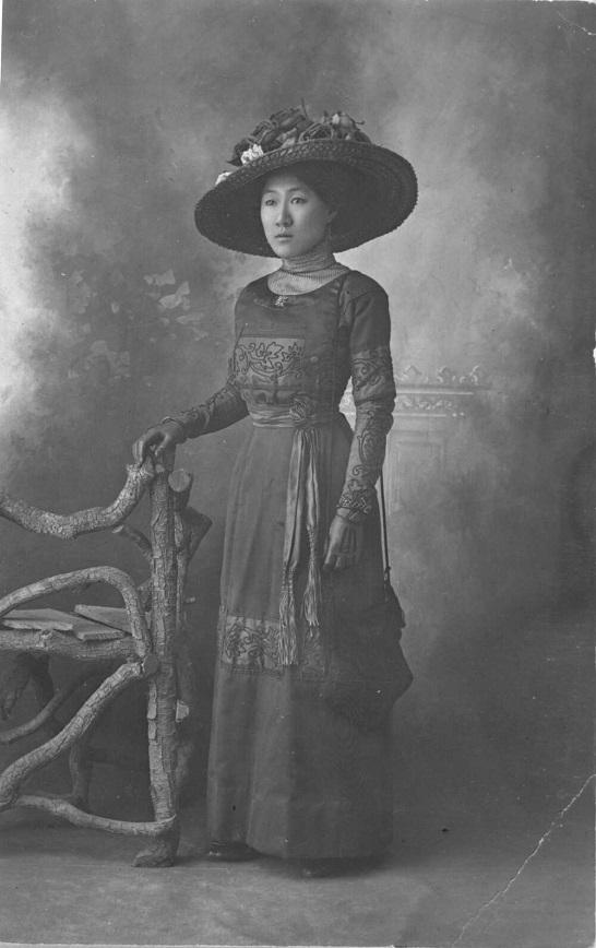 A black-and-white portrait of a woman wearing a long embroidered dress and a large hat adorned with flowers. She stands beside a rustic, wooden chair, gazing off-camera with a composed expression. 