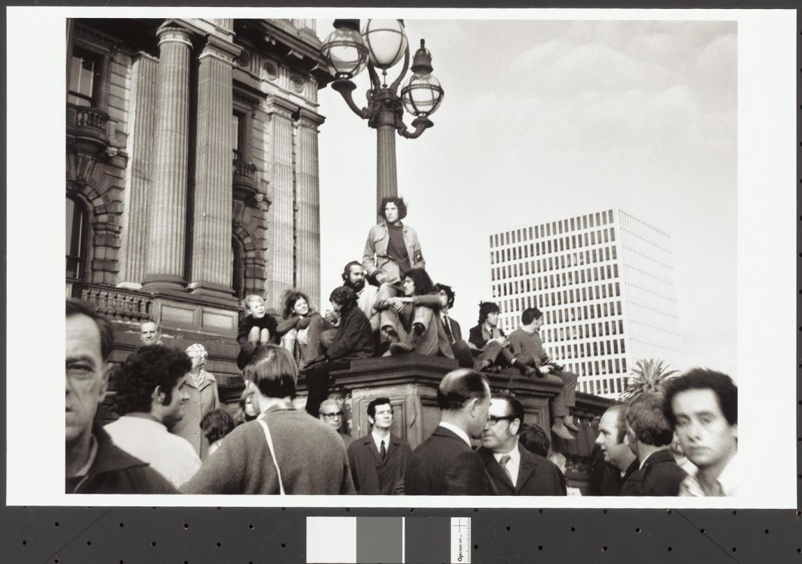 A black-and-white photograph capturing a Vietnam War moratorium protest in 1971 on the steps of the Victorian Parliament Building in Melbourne, Australia. The image shows a group of young protesters sitting on the base of a large lamp post outside the building, with some standing and others sitting closely together. The architectural columns of the parliament building loom behind them, while a modern office building is visible in the background. In the foreground are individuals, mostly men in suits.