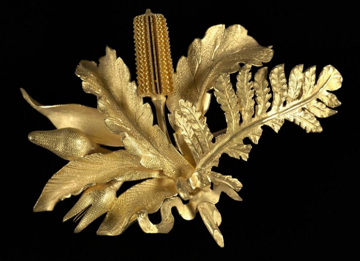 Ornate gold brooch shaped like native Australian flora, featuring detailed leaves, flowers, and textures against a black background.