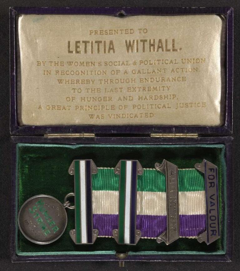 A medal displayed in a green velvet-lined case. The medal features a ribbon in purple, white, and green adorned with two silver bars. One bar is engraved with the date "AUG 1913," and another reads "FOR VALOUR." Accompanying the medal is a round silver disc engraved with "HUNGER STRIKE" and "WSPU" (Women's Social & Political Union). The lid of the case contains an inscription