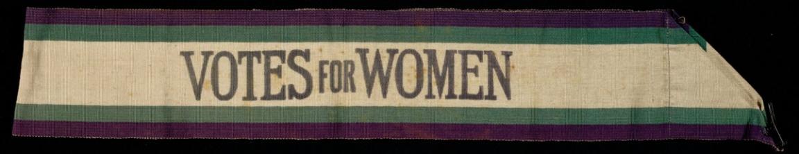An ivory-colored sash featuring bold black text that reads "Votes for Women," flanked by horizontal green and purple stripes. The fabric appears slightly worn, and the right end tapers into a pointed shape