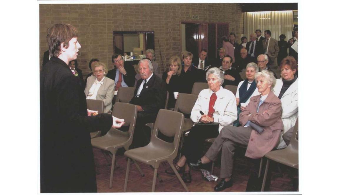 A photo of a woman standing and speaking to a room of seated and standing people.