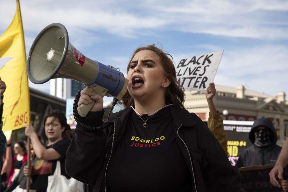 A person speaks into a megaphone during a protest rally, wearing a shirt with the text "Boorloo Justice." Behind them, others hold signs, including one that reads "Black Lives Matter." A yellow flag and buildings are visible in the background.