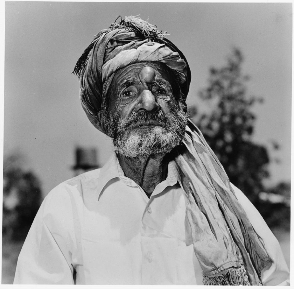 A black-and-white portrait of an older man with a weathered face and a beard, wearing a turban and a white shirt. He stands outdoors, with blurred trees in the background.