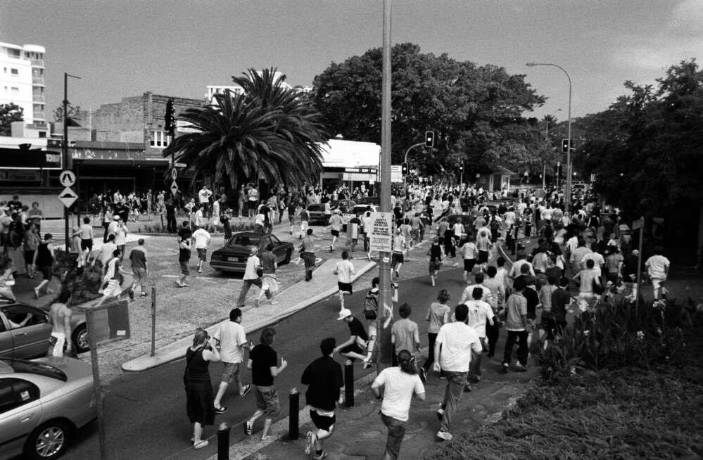 A large group of people, some running, fill a busy street near parked cars and a palm tree. The crowd moves toward a railway station. Buildings and traffic lights are visible in the background.