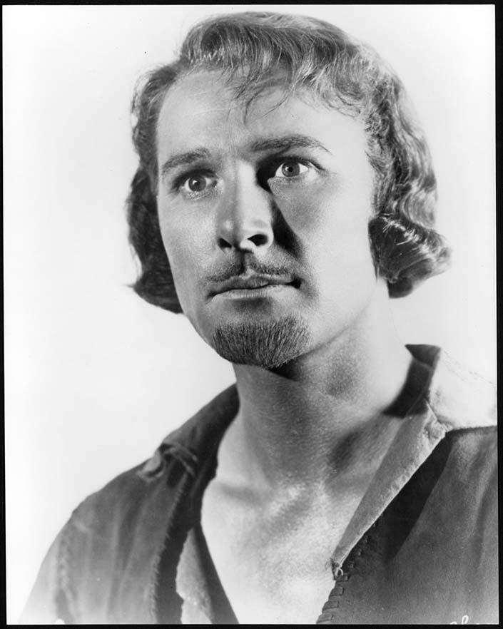 A black and white photograph of a man. He has an unbuttoned shirt showing his chest. He is staring off camera. He has a well groomed pencil moustache and a goatee. He has wavy blond hair.