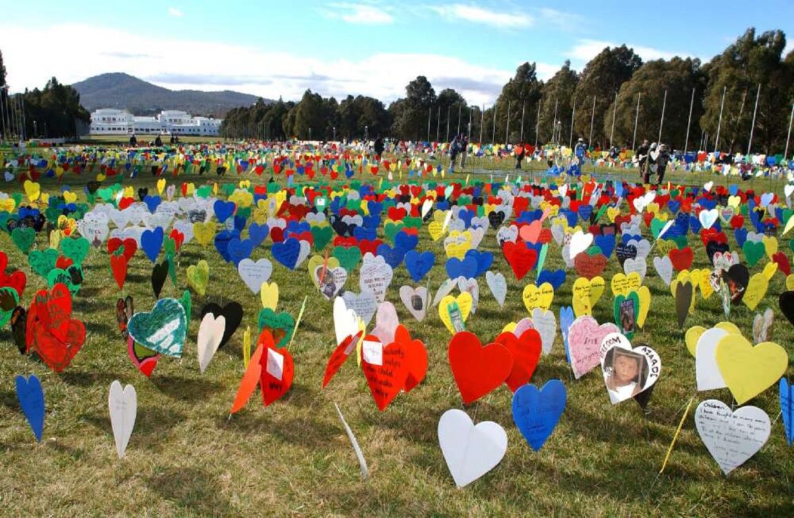 A large field filled with colorful heart-shaped signs, some with messages and images, displayed on the grass. Old Parliament House is visible in the background.