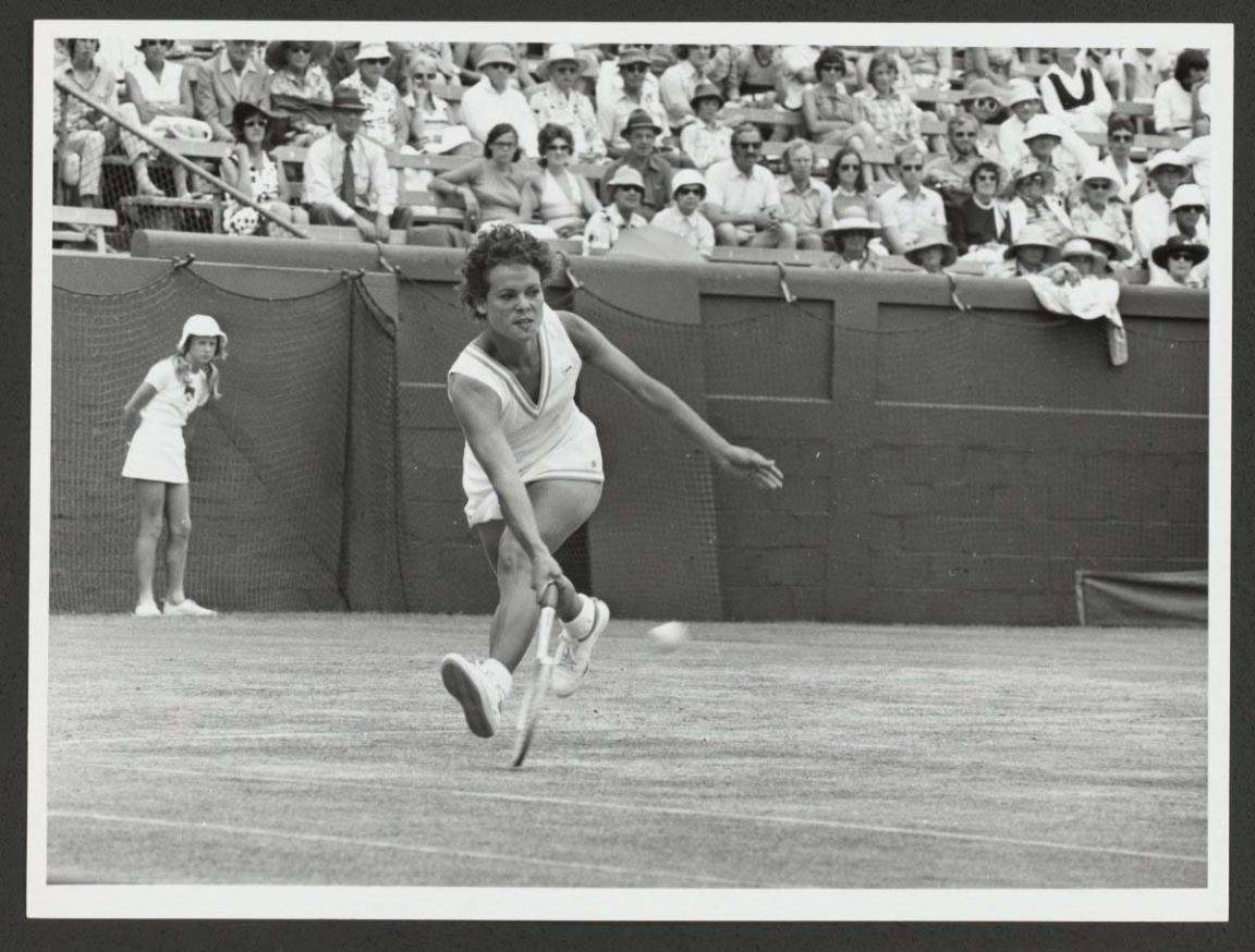 A black and white image of a woman wearing a white tennis dress lunging to hit a tennis ball. Behind her is a young woman in a white tennis dress and white bucket hat watching. There is a large crowd watching the game of tennis that is being played.