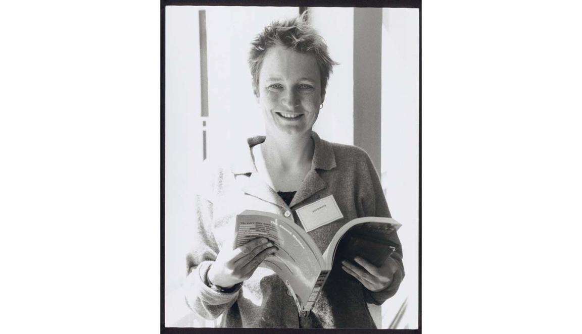 A black and white portrait photo of a young, freckle faced, smiling woman with very short, wild, fair hair, holding a book open.
