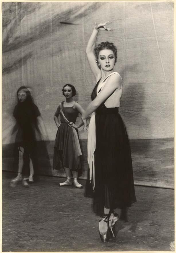 Ballerina Irina Baronova strikes a ballet pose for the camera. She is standing en point wearing a long black dress. Two dancers are watching her in the background