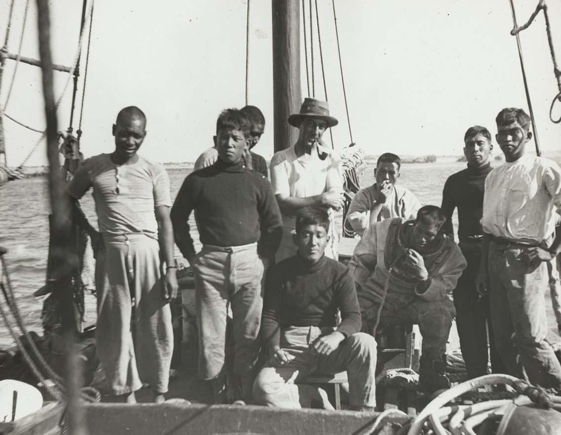 Nine men stand on the deck of a small boat. One is wearing a hat, the others are not. Three men are sitting. In the background the ocean can be seen.