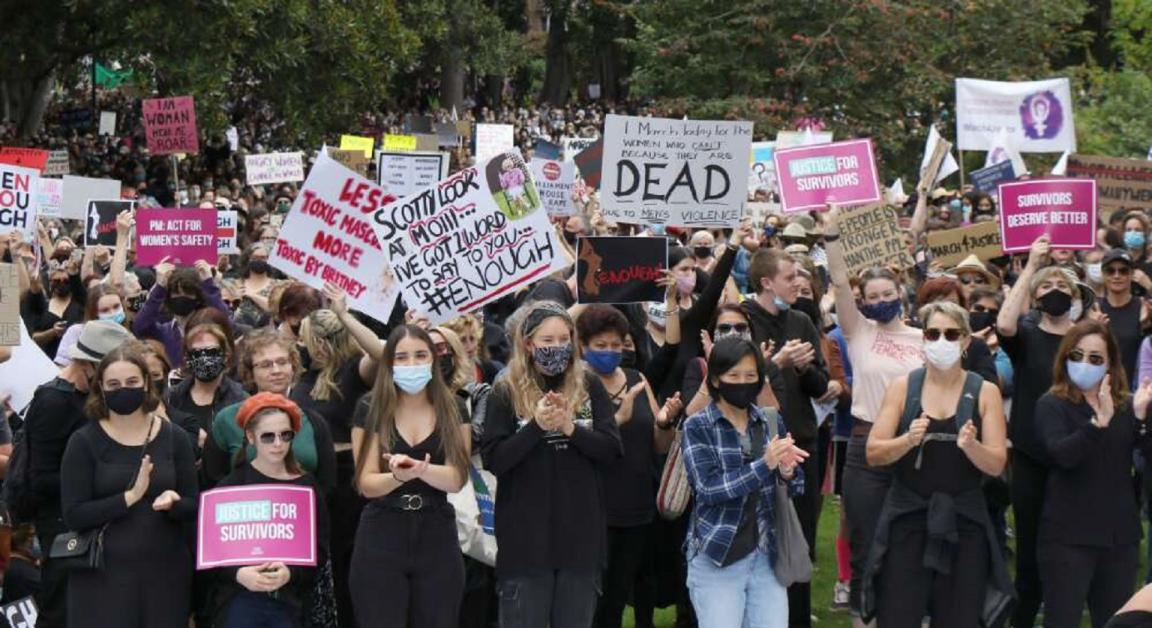 A large crowd of demonstrators gathered outdoors, many wearing masks and holding signs with messages such as "Justice for Survivors," "Survivors Deserve Better," and "Enough." Some individuals clap while others raise placards. Trees line the background.