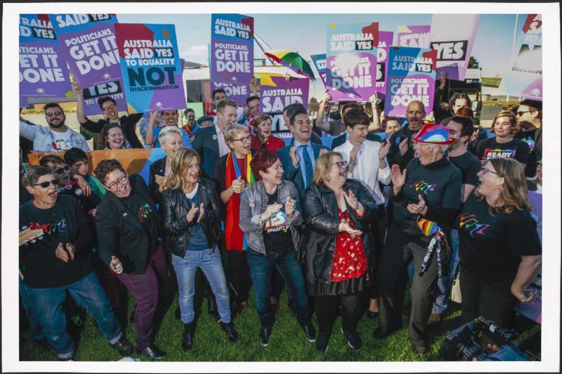 A group of people gathered on a lawn, cheering and smiling, holding signs with slogans such as "Australia Said Yes" and "Politicians Get It Done." Several individuals at the front are clapping and wearing colorful clothing. The crowd appears jubilant, celebrating together outdoors.