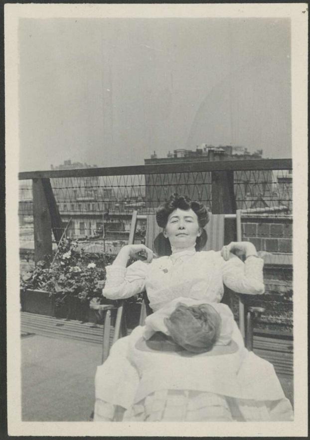 A black-and-white photograph of a woman reclining on a rooftop, dressed in a light-coloured Victorian-era dress, hands resting behind her head.