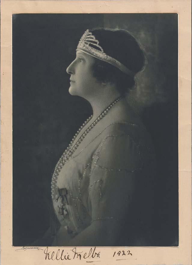 A formal portrait photograph of a lady wearing a diamond tiara. She is in profile. She has short hair and a lace gown on. She is wearing a long string of pearls. The photograph is signed 'Nellie Melba 1922'