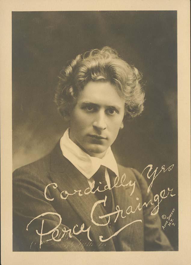 A sepia toned portrait of a young man with wavy blond hair. He is wearing a shirt with large lapels and black tie. Signed on the photo are the words 'Cordially yours, Percy Grainger'