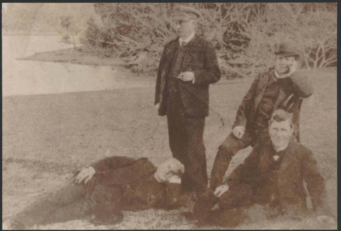 A black and white photograph of three men in suits and waistcoats. One man is standing another is sitting on a chair, another is sitting on the grass and the fourth is laying at the feet of the standing man. They are outside and positioned in front a some large bushes.