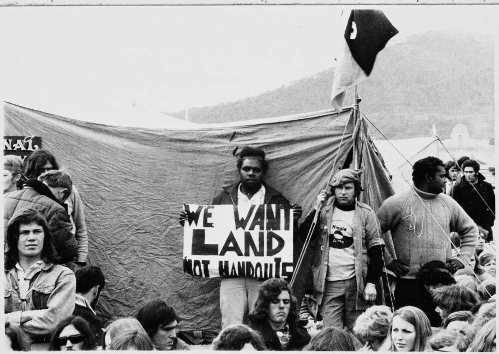 A group of people gathered in front of a tent, with one person holding a sign that reads "We want land not handouts" at a land rights demonstration. A flag is visible in the background. 