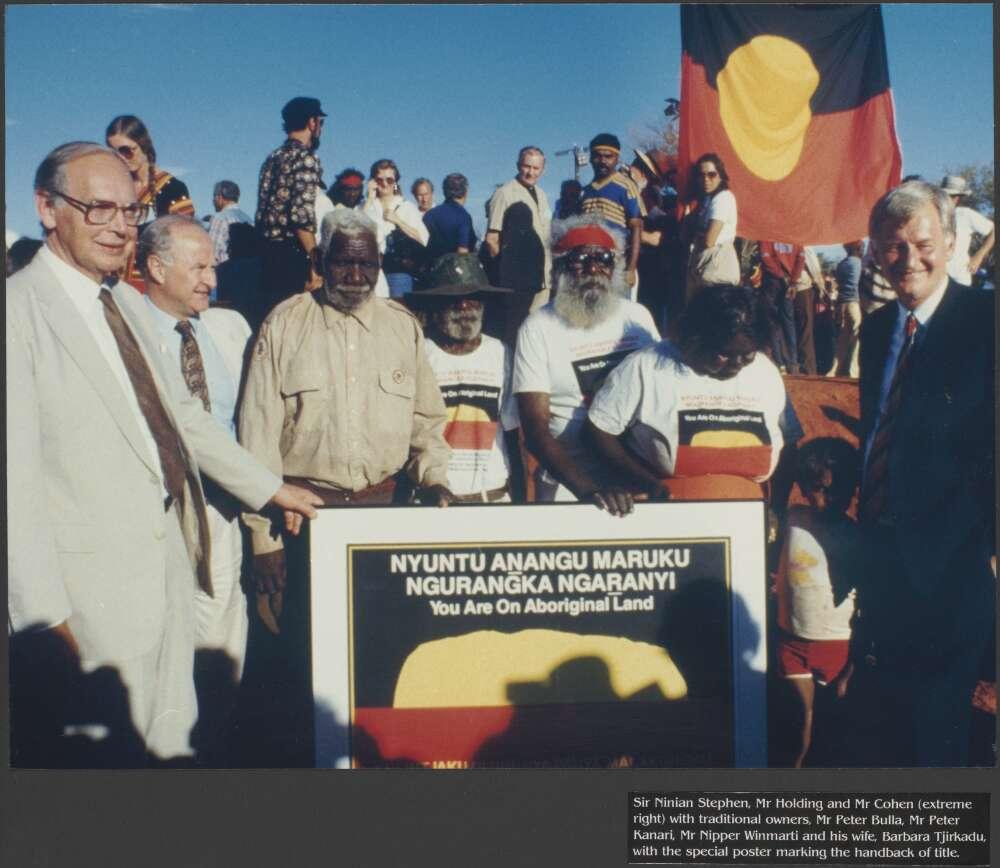  A group of people standing outdoors, holding a poster that reads "You Are On Aboriginal Land," with the Aboriginal flag prominently displayed. Several individuals are wearing shirts with similar designs, and others in the background observe the scene. An Aboriginal flag is raised in the background.