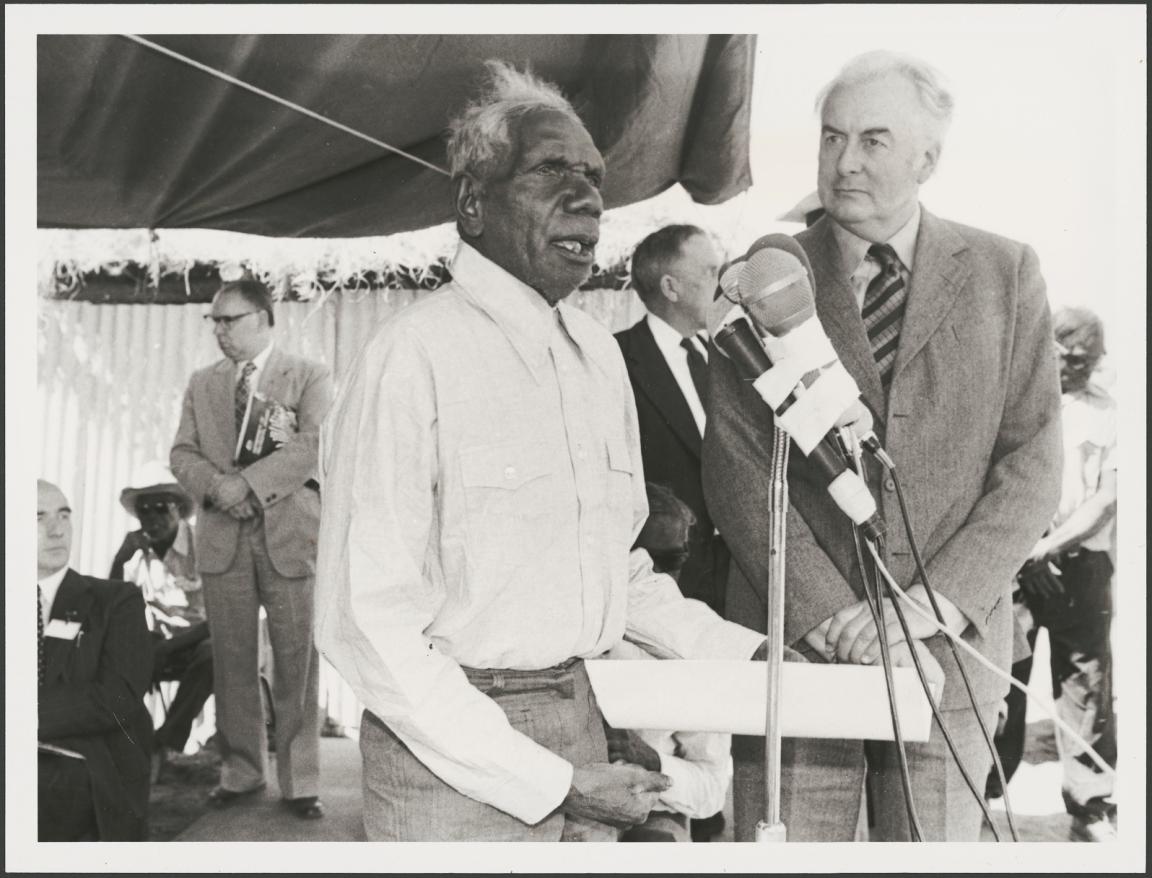 Vincent Lingiari addresses a crowd at a microphone while Gough Whitlam stands beside him. Several people are visible in the background, observing.