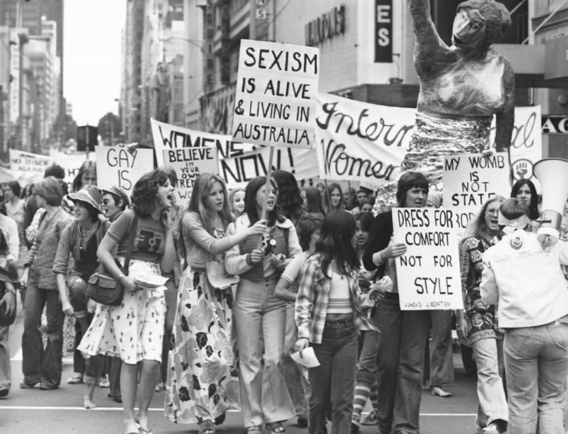 A group of women marches down a city street, holding various protest signs. Signs include messages like "Sexism is alive & living in Australia" and "Dress for comfort, not for style." A large sculpture is visible in the background.