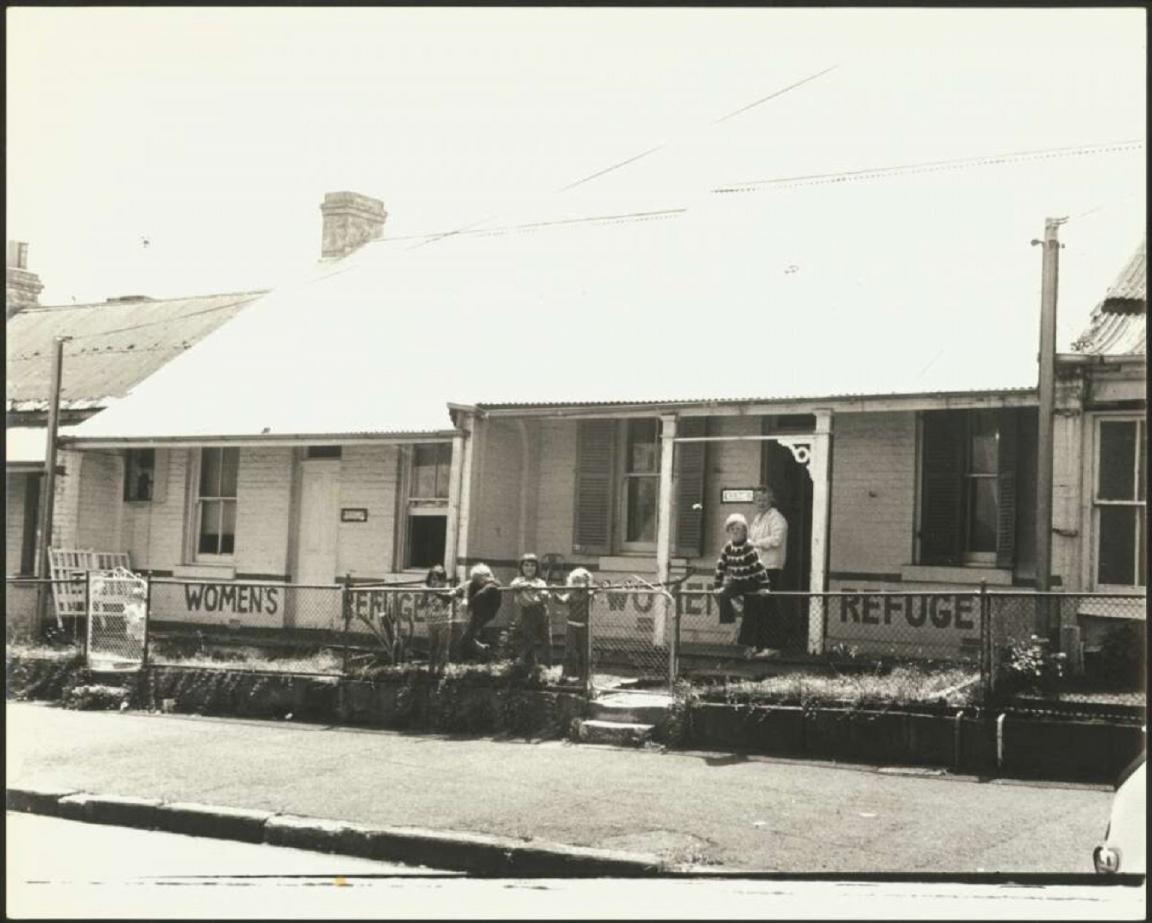 A black-and-white photograph of six children playing in the front yard of the Elsie Women's Refuge, supervised by a woman standing near the front door. The building is a simple single-story structure with a sign reading "Women's Refuge" on the fence.