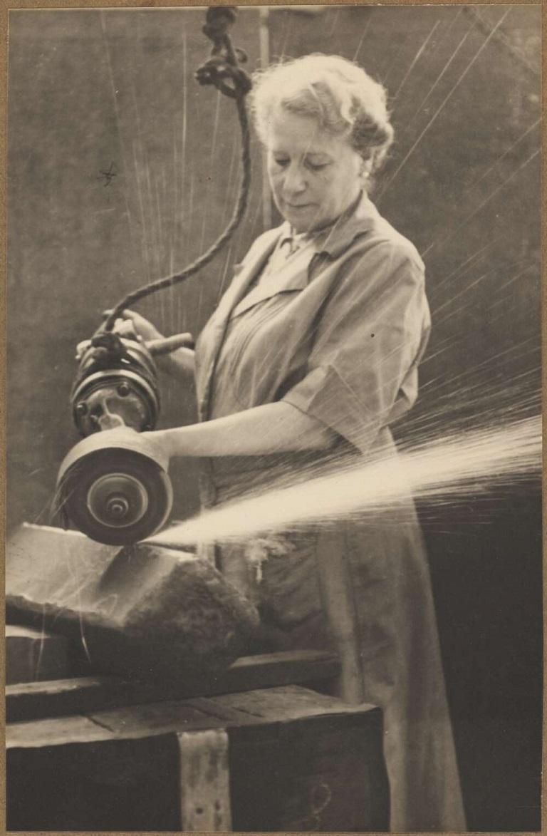 A black-and-white photograph of a woman in work attire using a grinding tool on a billet of armor-piercing steel, with sparks flying from the object being ground.