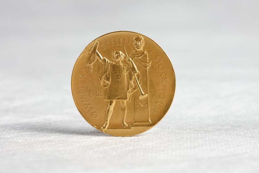 A gold medal standing upright on a white cotton surface. The medal shows a figure holding a trumped and a wine amphorae. Behind the figure is a bust on a stand. In raised lettering around the edge of the medal are the words 'Olympiska Spelen Stockholm' The year '1912' is also present.