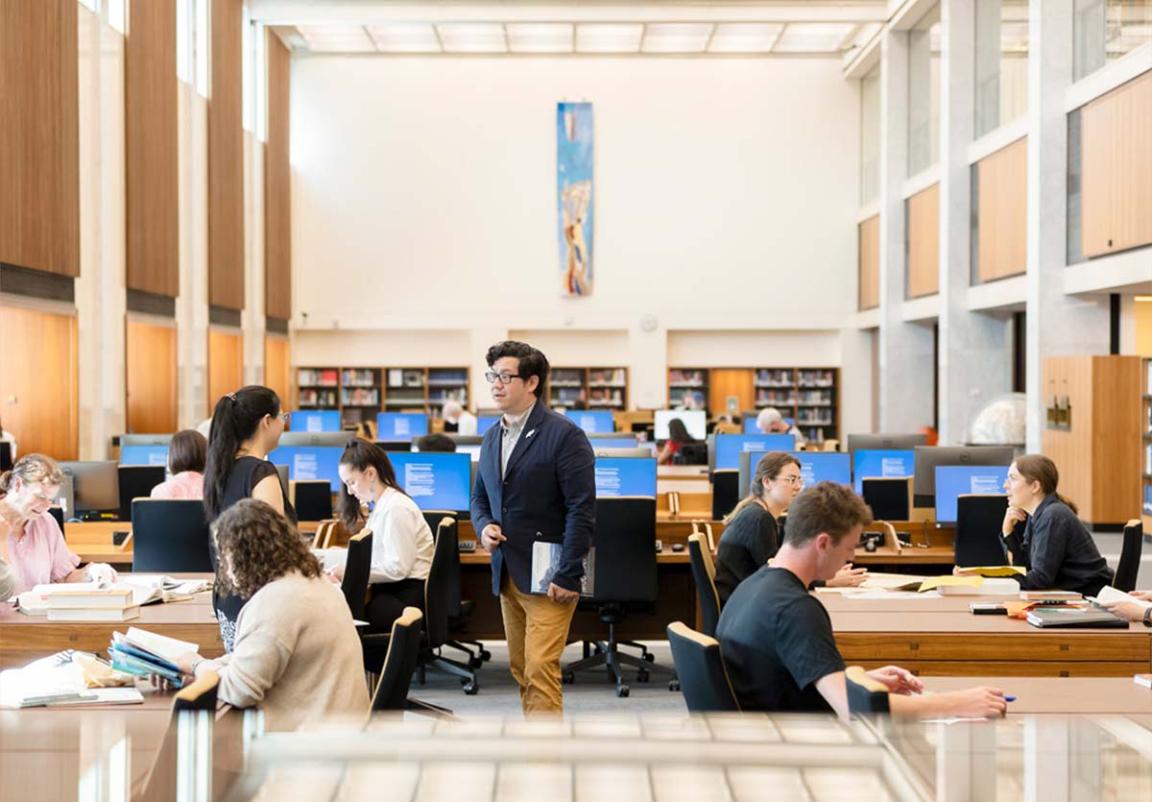 A man talks to a woman in the reading room of the National Library of Australia while other people work at tables and computers