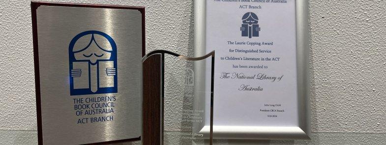 A silver and wooden plaque, glass trophy and framed certificate for the 2024 CBCA Laurie Copping Award given to the National Library