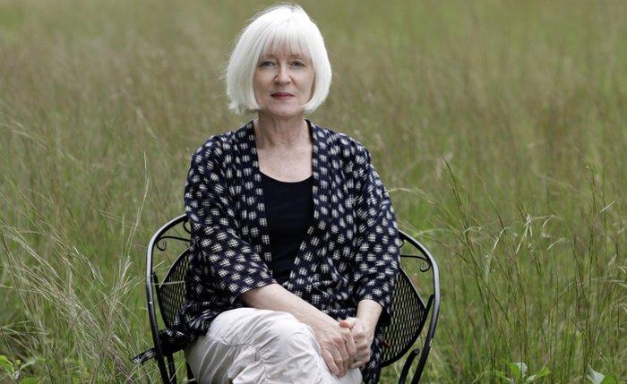 Dr Thompson has short white hair and sits outdoors in a grassy field on a black metal chair. She is wearing a dark, patterned shawl over a black top and light-coloured pants. Her hands are clasped in her lap, and she has a calm, neutral expression, gazing directly at the camera. The background is filled with tall green grass, creating a serene, natural setting.