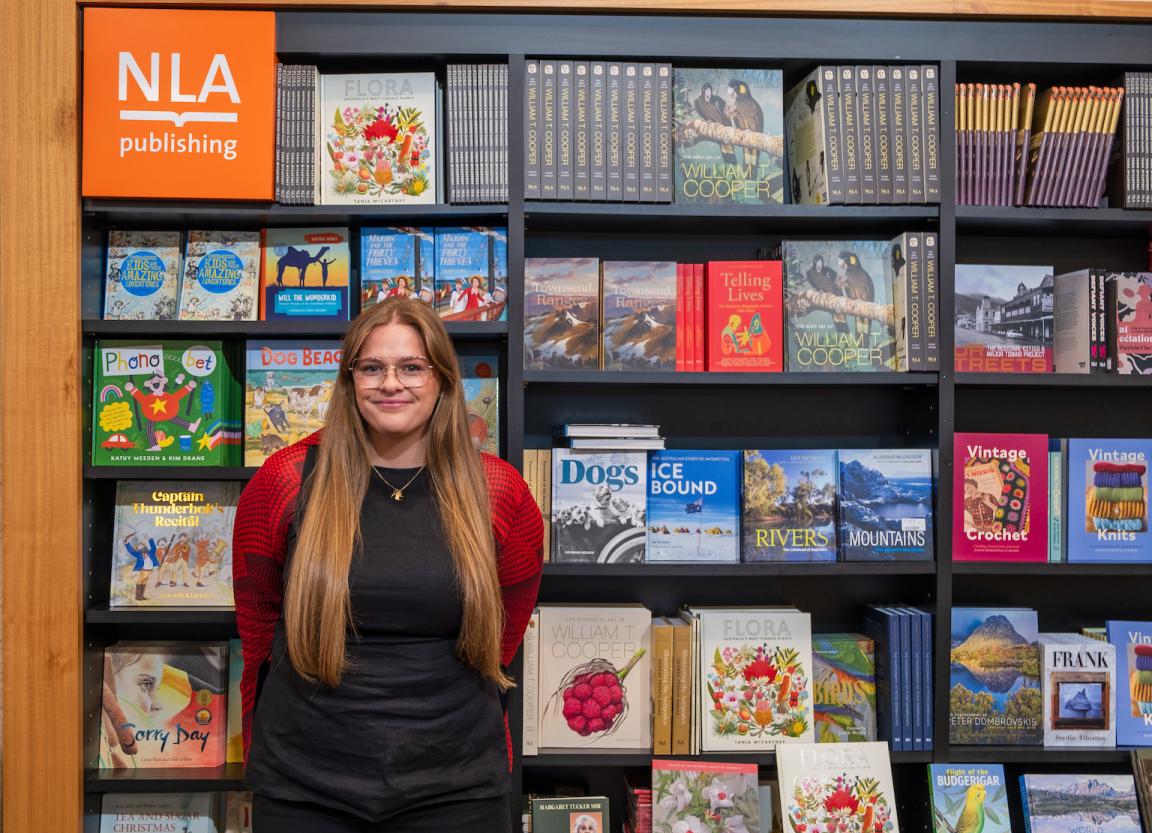 Young woman, Maddie from NLA Publishing, standing in front of a wall of NLA Publishing books in the Bookshop