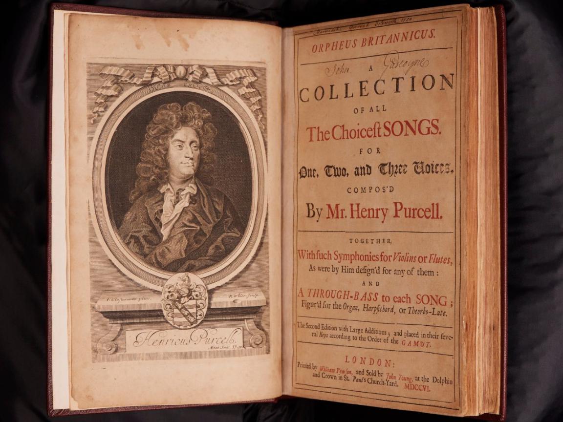 Old book sitting open on a black cushion. On the left page is a portrait of a man with large rolled hair, on the right is the title page with text reading 'Orpheus Britannicus; A collection of all the choicest songs for one, two and three voices, composed by Mr. Henry Purcell'