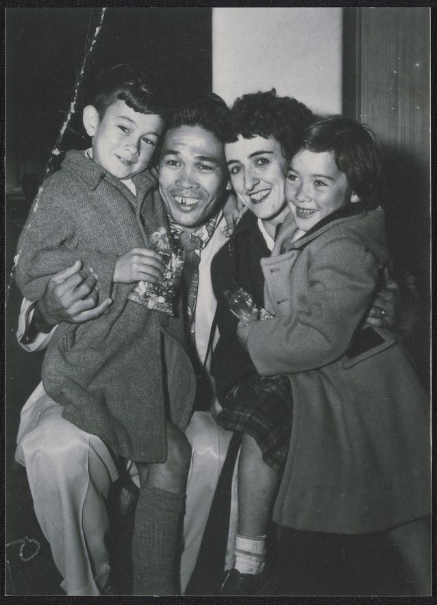 A man seated in the center, holding a young child on each side. A woman stands beside him, leaning close. The children wear coats, and all four individuals are smiling.