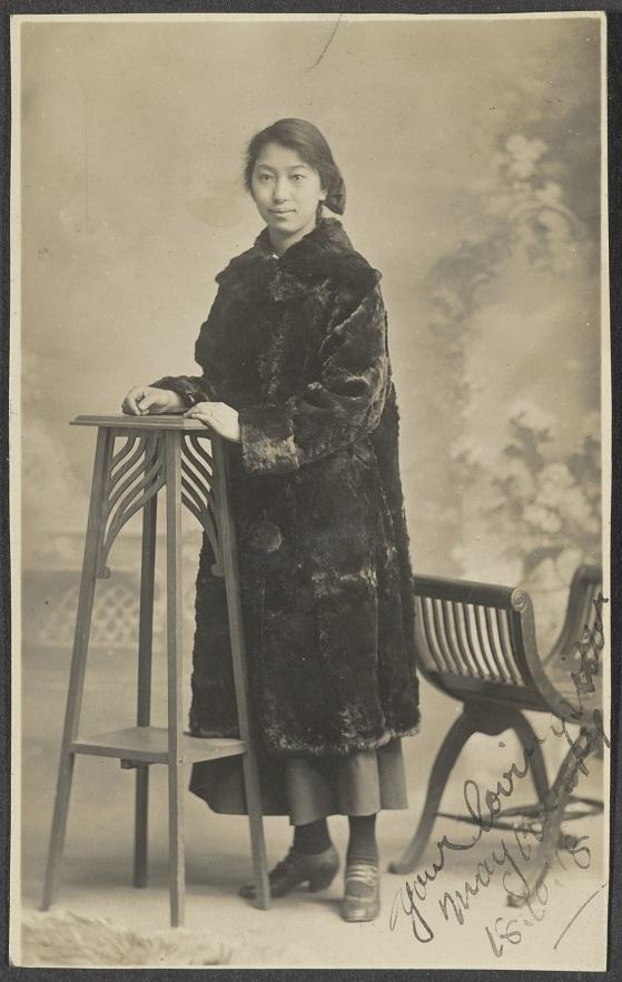Kwong Mui Ying (May) is standing in a studio setting, dressed in a long fur coat over a modest skirt, with one hand resting on a decorative wooden stand and the other by her side. A vintage chair is positioned behind her, and the backdrop features a painted garden scene. The photograph includes an inscription in the lower-right corner.