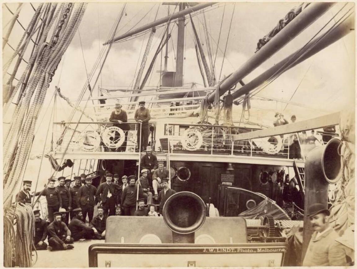 A sepia-toned photograph showing the deck of a large sailing ship with a group of sailors and officers posing for the camera. The crew members, dressed in uniforms and maritime attire, are gathered across different levels of the ship, with some standing on the upper deck and others grouped on the main deck. The ship's rigging, lifebuoys labeled "City Line Glasgow," and other maritime equipment are prominently visible.