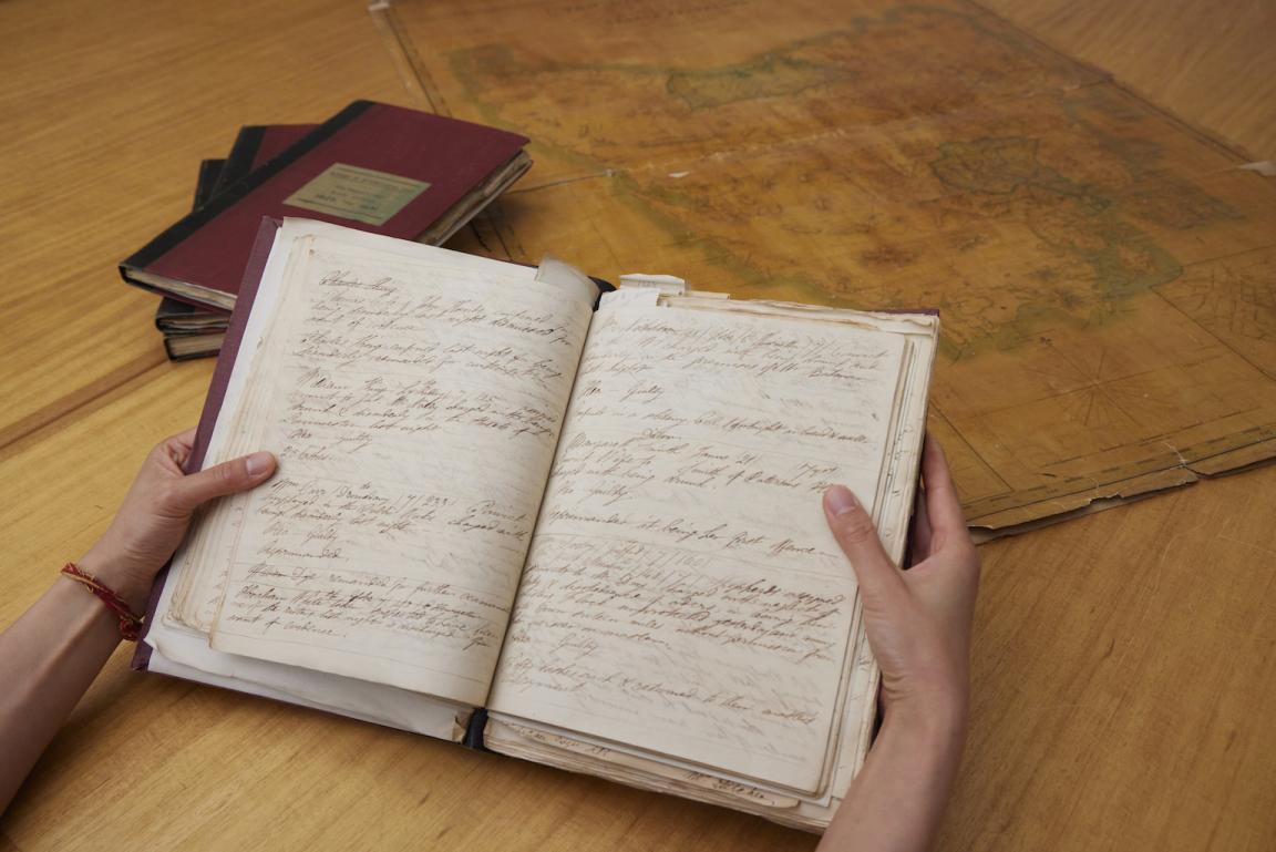 Hand holding an old notebook with handwritten text. In the background is a stack of books and an old map.