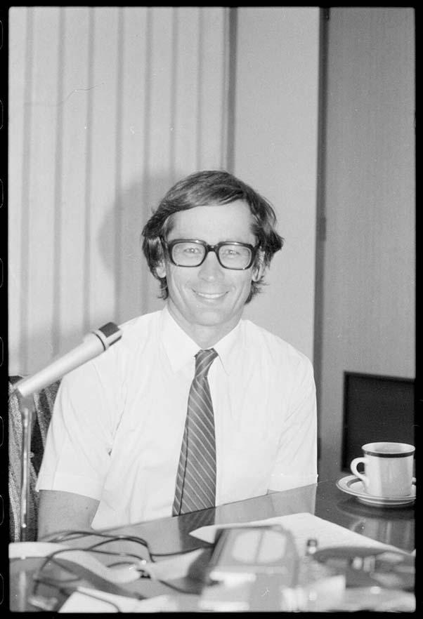 black and white photo of Dick Smith sitting at a table with a microphone set up on his right and a cup on his left
