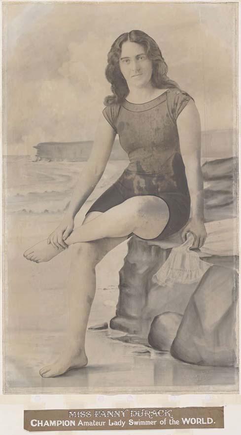 A sepia toned photograph of a woman sitting on a rock. She is wearing an old fashioned swim suit. She is sitting crossed legged. Beneath her is a caption saying 'MISS FANNY DURACK' Champion Amateur Lady Swimmer of the World'