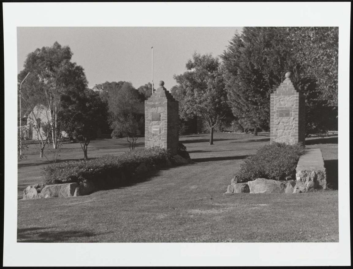 A black and white photograph of the main garrison gates of the former Cowra Prisoner of War camp. The gates now sit in a grassy park surrounded by trees and bushes.