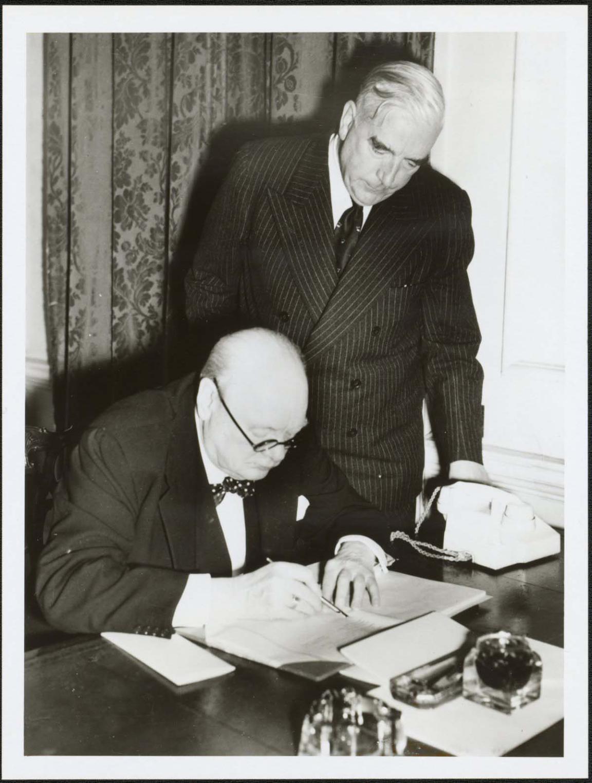 British Prime Minister Winston Churchill is seated at a table wearing glasses. He is signing a document. Standing behind him and leaning over his shoulder is Australian Prime Minister Robert Menzies. Both men are wear dark suits with white shirts. Menzies is wearing a dark tie, Churchill a bow tie.