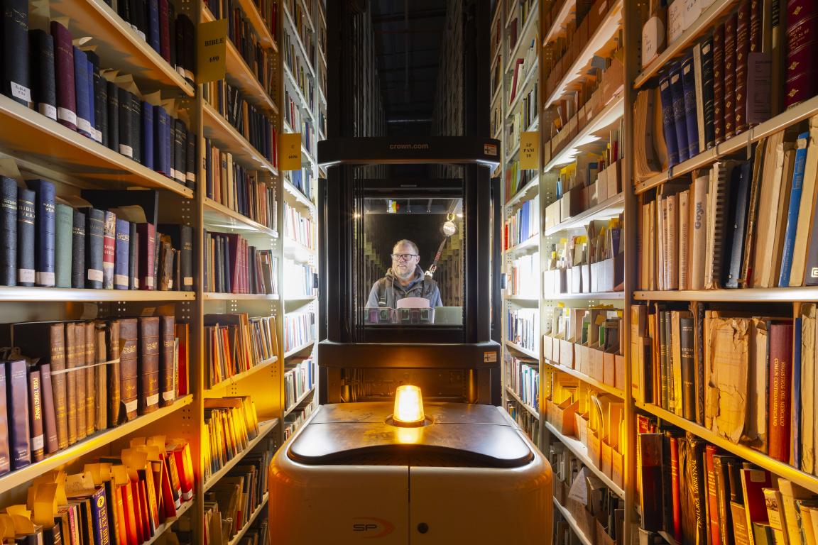 Person operating a lift between tall shelves filled with books, with a bright light at the front of the lift and warm overhead lighting.