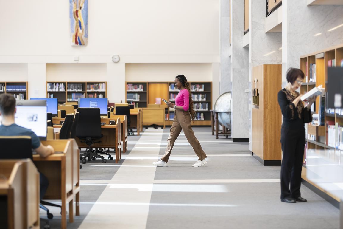 A person in a bright pink top and beige pants walks through an aisle in the Main Reading Room at the Library. People are sitting at wooden desks working on computers, and there are shelves filled with books and a large globe around the edges of the room. To the right, another person stands near a bookshelf, reading a book.