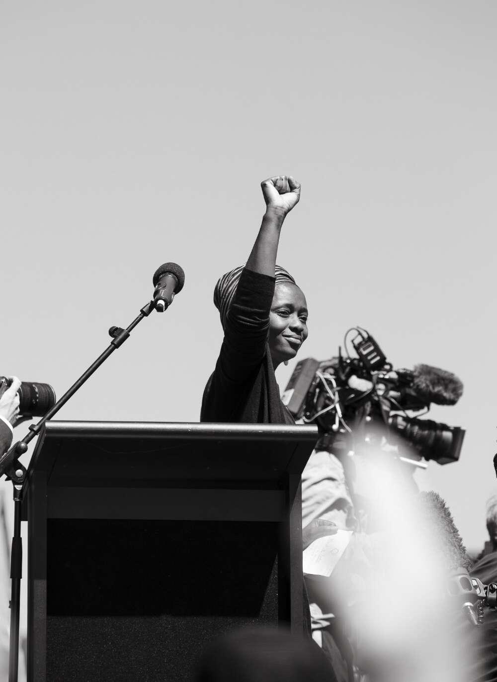 Aminata Conteh-Biger, wearing a headscarf, stands at a podium with a raised fist, captured in black and white. Cameras and microphones surround Aminata, capturing the scene.