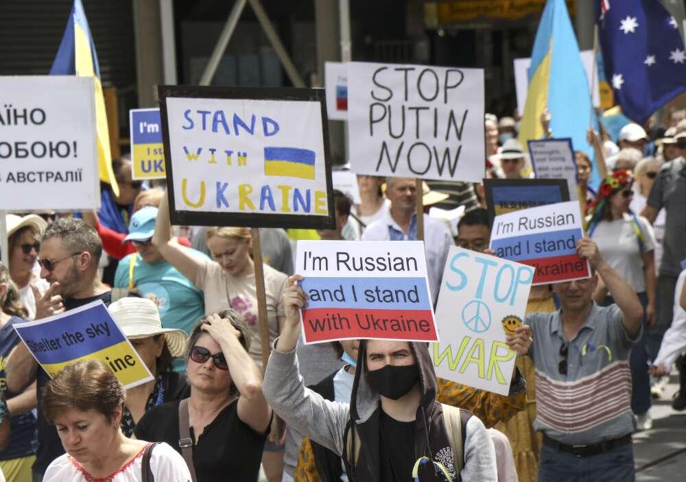 Crowd of protesters holding signs with messages like 'Stand with Ukraine,' 'Stop Putin Now,' and 'I'm Russian and I stand with Ukraine.' People wear a mix of hats and sunglasses, with Ukrainian flags visible in the background.