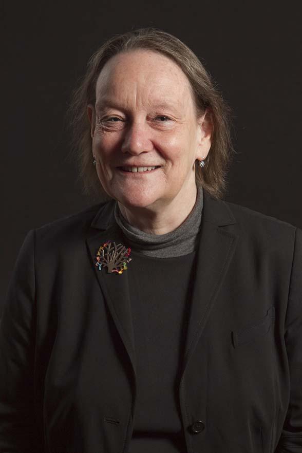 A portrait of a smiling woman wearing a black cardigan over a black top and a grey sweater. She is also wearing a colourful brooch shaped like a tree with colourful leaves.
